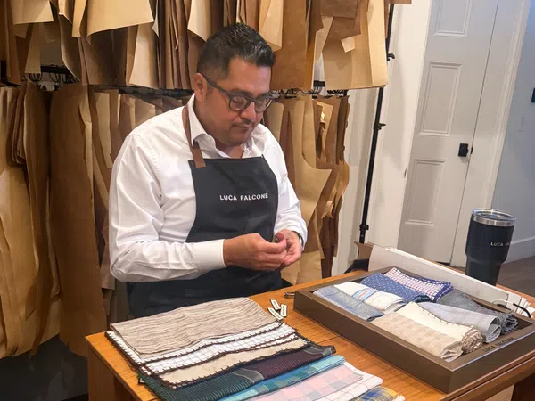 Tailor wearing Luca Falcone apron working with fabric samples in a workshop filled with hanging patterns.