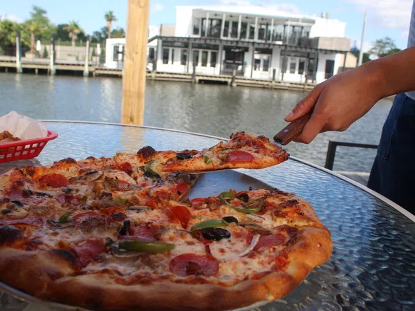 Person lifting a slice of pepperoni pizza from a glass table by the waterfront with a house in the background