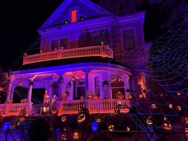Large Victorian house decorated with glowing jack-o'-lanterns, skeletons, giant spiders, and web lights for Halloween night.