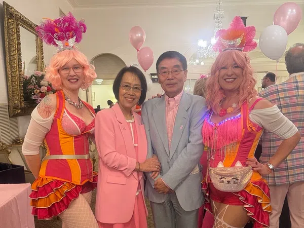 Two women in colorful circus costumes and pink wigs pose with a couple dressed in pastel pink and gray at a festive event.