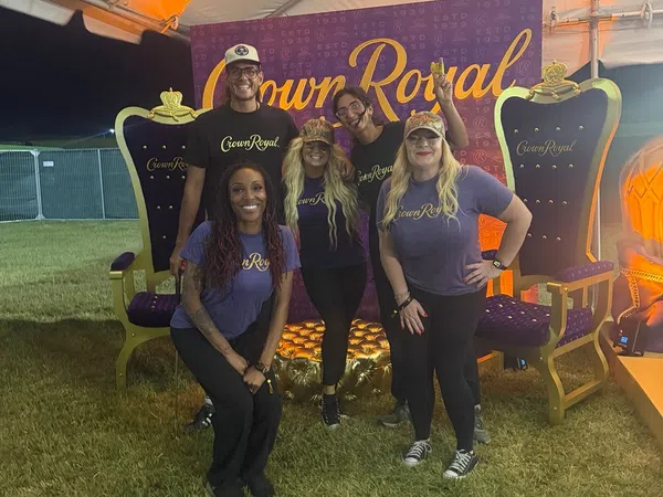 Five people wearing Crown Royal shirts pose in front of a Crown Royal backdrop between two large decorative royal chairs.