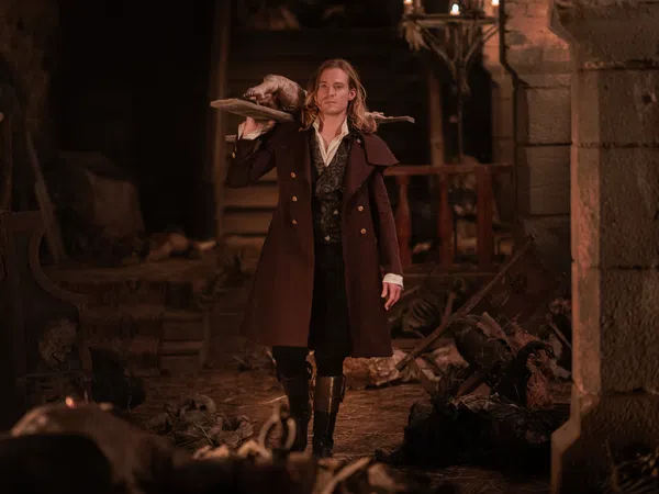 Man in historical coat carrying a wooden board with a dead animal in a dimly lit stone room with skeletons.