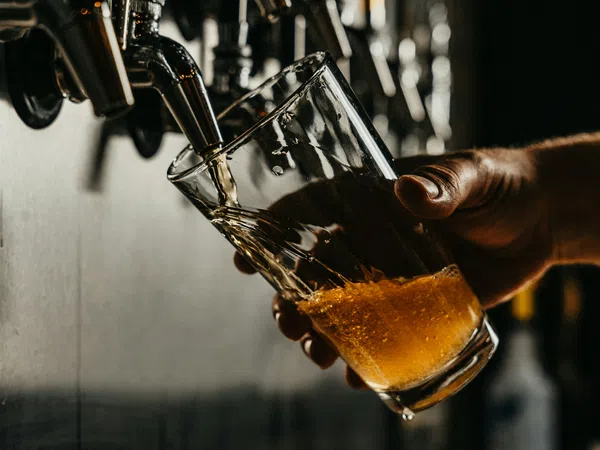 Hand pouring amber beer from tap into tilted glass held by hand in dimly lit bar setting.