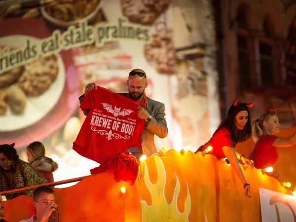 Man holding a red Krewe of Boo t-shirt on illuminated Halloween parade float with people in costumes.