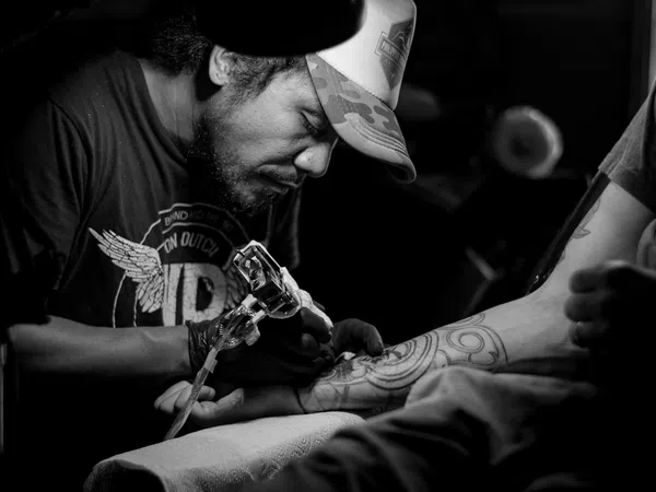 Black and white photo of a tattoo artist focused on inking a geometric tattoo on a client's forearm in dim light.