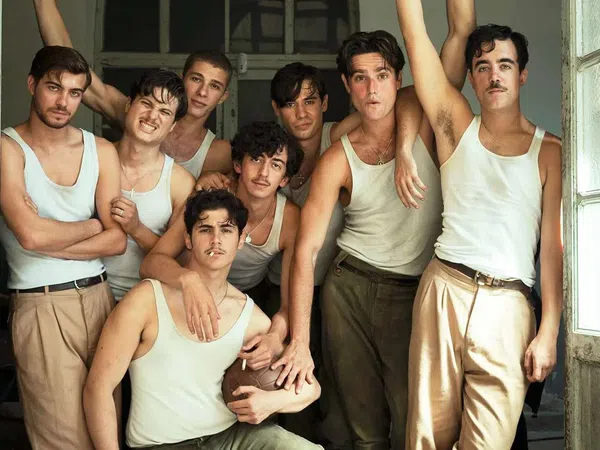Group of eight young men wearing white tank tops and beige pants posing closely in a vintage room near open windows