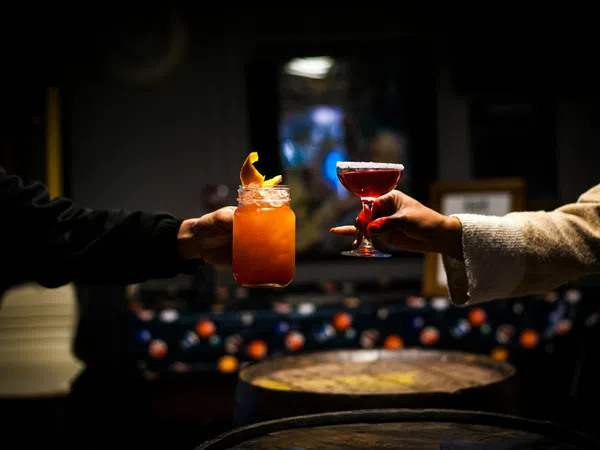 Two hands holding up colorful cocktails in dimly lit bar with wooden barrels in the foreground.