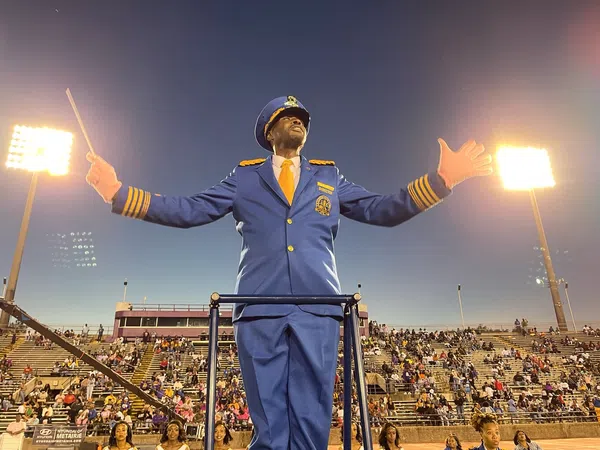 Band director in blue uniform conducting with baton at a stadium filled with spectators during evening.