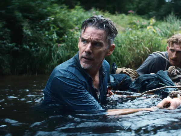 Two men cautiously wading through a river surrounded by dense greenery, holding onto a rope for support.