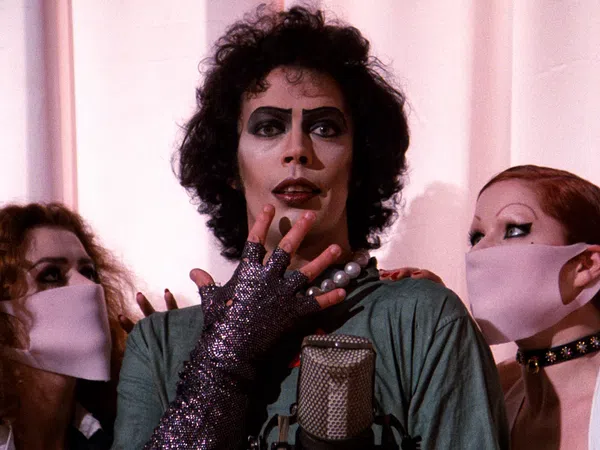 Actor in iconic glam rock makeup and costume with two masked fans on either side against a pale curtain.