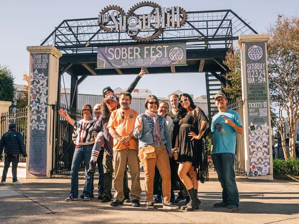 Group of people posing and smiling under Sober Fest banner at outdoor event entrance on sunny day
