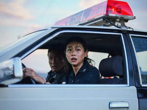 Female police officer and male colleague in a police car with red siren light, looking concerned through open window.