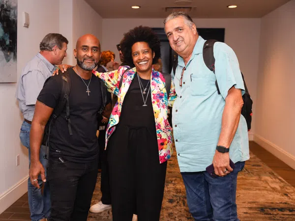 Three diverse adults smiling and posing together indoors at a social event in a hallway.