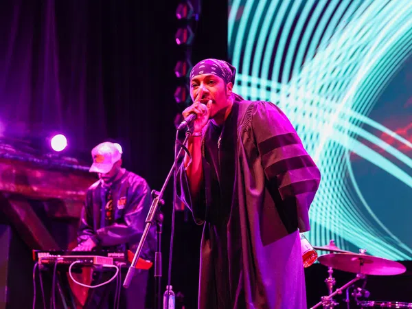 Hip-hop artist performing on stage with microphone and DJ in the background under vibrant purple and blue lights.