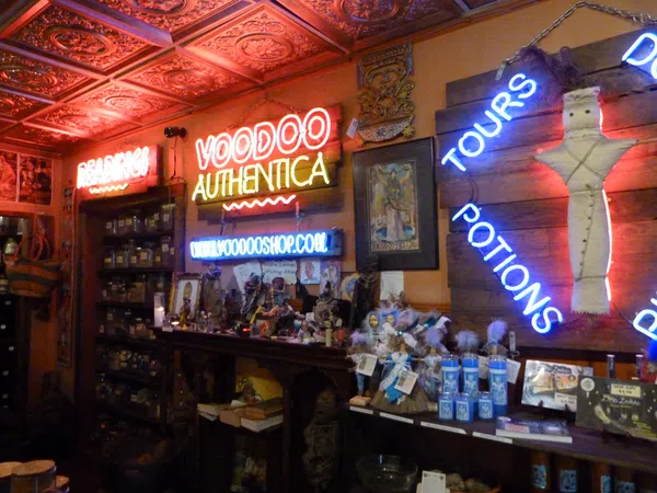 Interior of a voodoo shop with neon signs, shelves of jars, potions, dolls, and mystical items under ornate ceiling tiles.