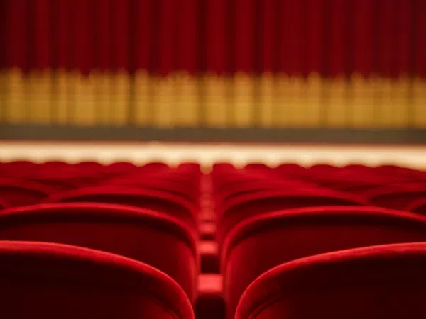 Empty red theater seats facing a closed stage curtain with golden accents under warm lighting.