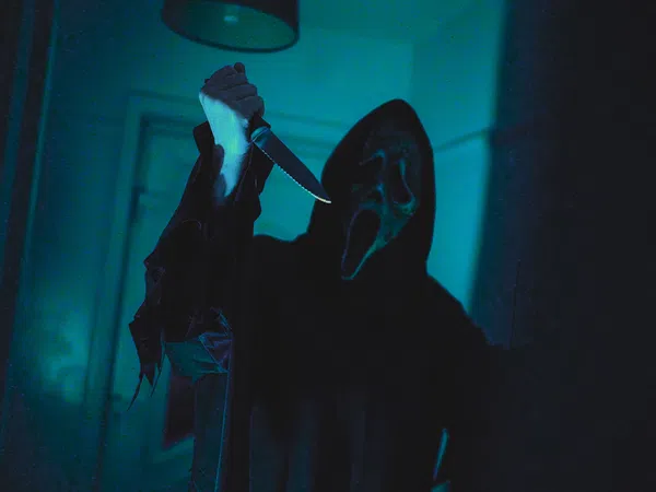 Person wearing a ghost face mask and dark robe holding a raised knife in a dimly lit room.