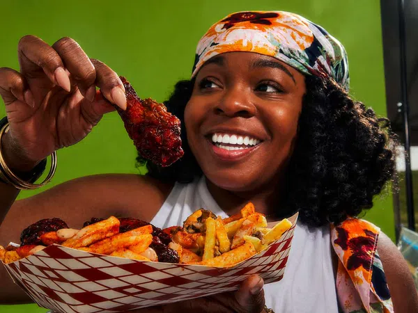 Smiling woman holding a saucy chicken wing and a tray of seasoned fries against a green background.