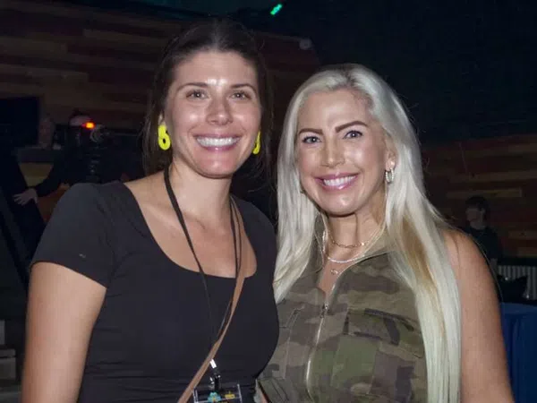 Two smiling women posing indoors, one with long blonde hair in camouflage top, the other with dark hair and yellow earrings.