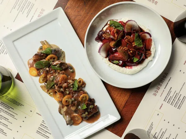 Gourmet dishes with sauces and herbs served on white plates surrounded by menus and wine glasses on wooden table.
