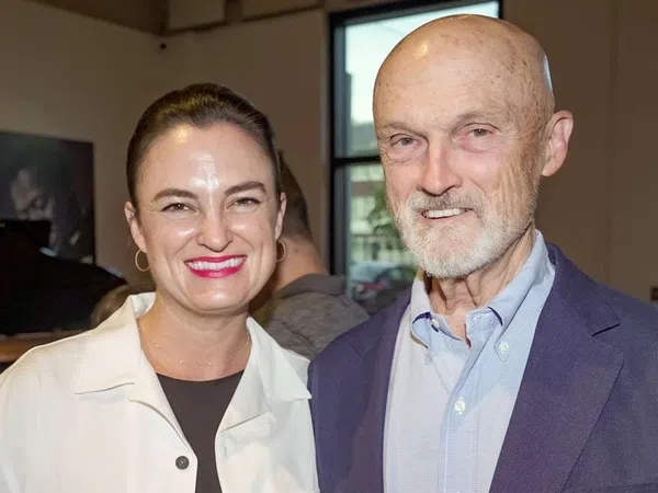 Smiling woman and older man standing indoors near a window in a casual social setting.