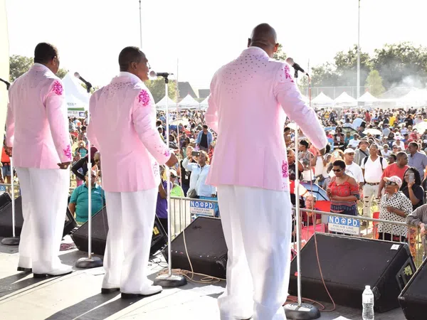 Five male singers in pink jackets performing on outdoor stage before large audience at daytime event.