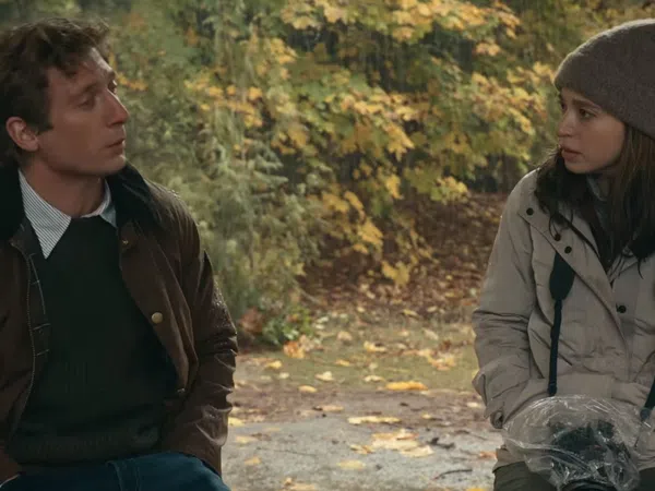 Man and woman dressed in autumn clothing sit outdoors surrounded by fall foliage during a conversation.
