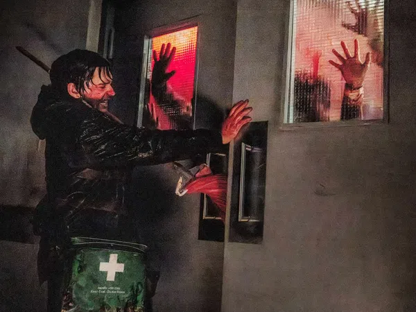 Man pushes hospital doors with bloody hands as silhouettes press against red-lit windows in a tense scene.