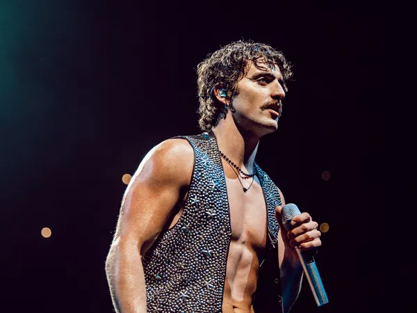 Muscular male singer with curly hair and sleeveless studded vest performing on stage with microphone