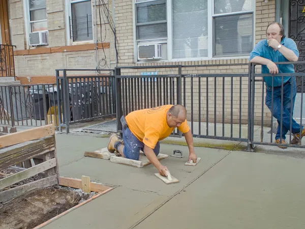 Construction worker smoothing fresh concrete on a sidewalk while another man watches at a residential building.