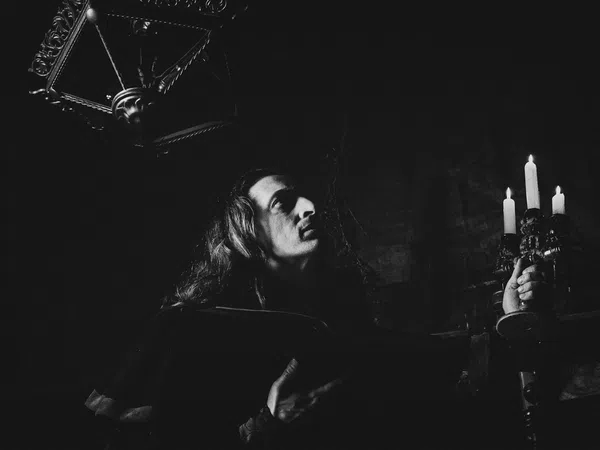 Black and white photo of a person holding a candelabra with burning candles in a dark, gothic setting.