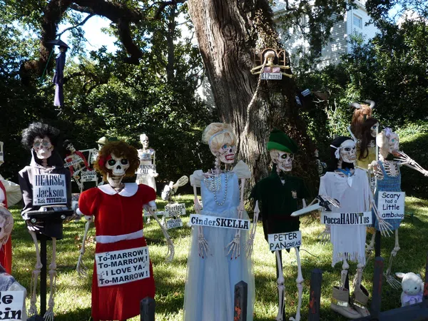 Halloween skeleton characters with humorous signs in a sunlit yard with trees and a house in the background.