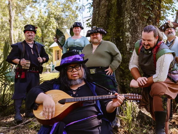 Group of six people in medieval and pirate costumes posing outdoors with a lute player in front under a large tree.