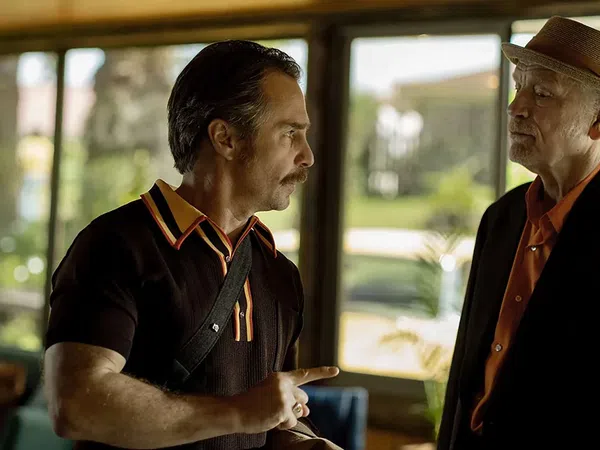 Two men in conversation, one wearing a hat and suit, the other in a black shirt with orange collar details indoors near windows.