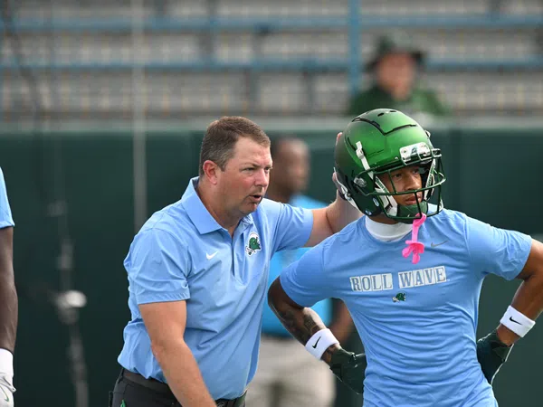 Football coach instructing player wearing green helmet and blue Roll Wave shirt during practice session