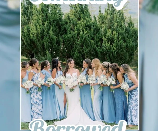 Bride in white dress surrounded by bridesmaids in blue dresses holding bouquets outdoors with greenery background