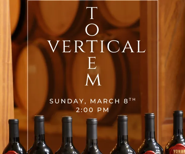 Seven wine bottles with Yonah Mountain Vineyards Totem labels lined up in front of wooden barrels for a vertical tasting event.