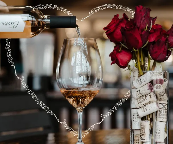Wine being poured into a glass with red roses in a cork-filled vase on a wooden table for Valentine's Day.
