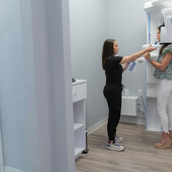 Dental technician assists patient in a floral shirt and white pants during a panoramic X-ray scan in a clinic.