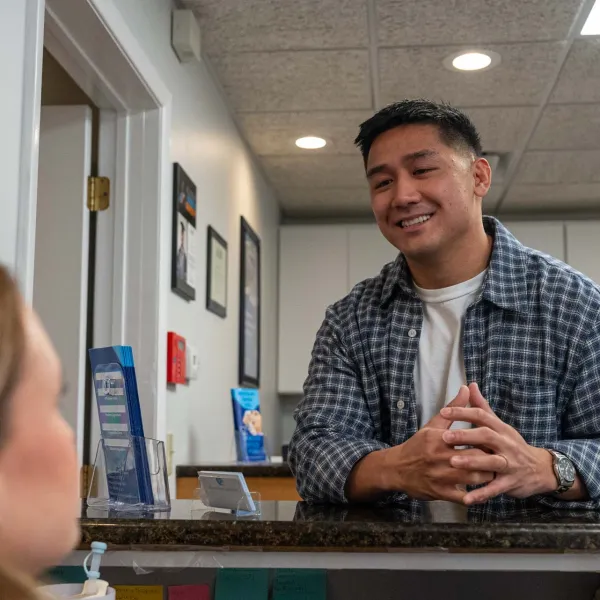 Smiling man in plaid shirt talks to a woman across a reception desk in a professional office setting.