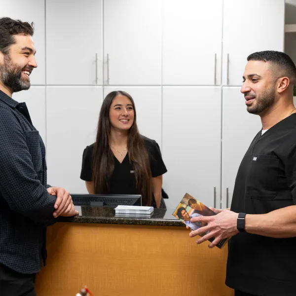 Two men speaking at a dental clinic reception desk with a female staff member smiling behind the counter