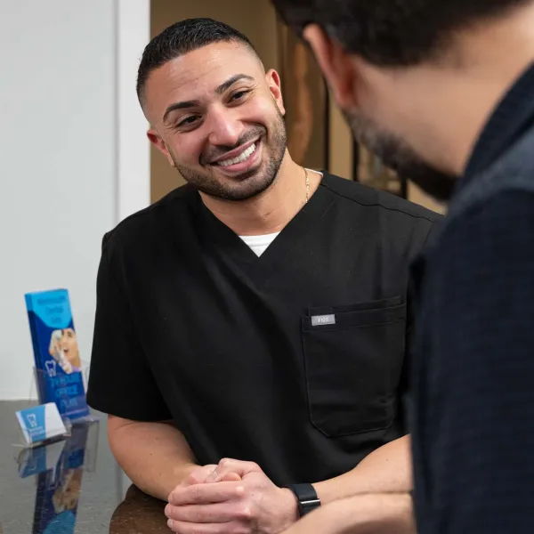 Smiling male healthcare professional in black scrubs talking with a patient across a desk in a medical office.