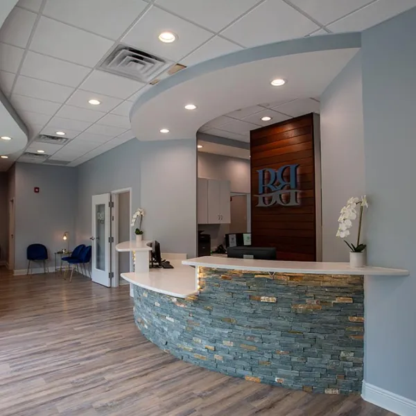 Modern dental office reception area with curved stone desk, wood flooring, blue chairs, and soft lighting.