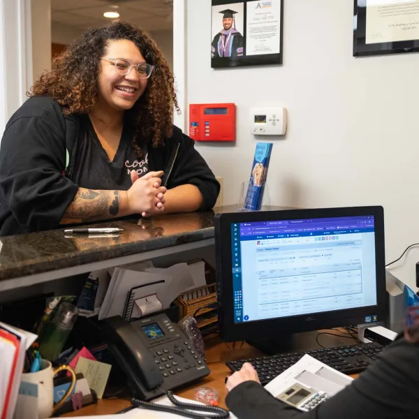 Smiling woman with curly hair talks to receptionist at office counter with computer and phone.