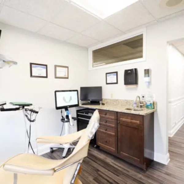 Bright dental office with beige patient chair, monitors, cabinets, and certificates on the wall.