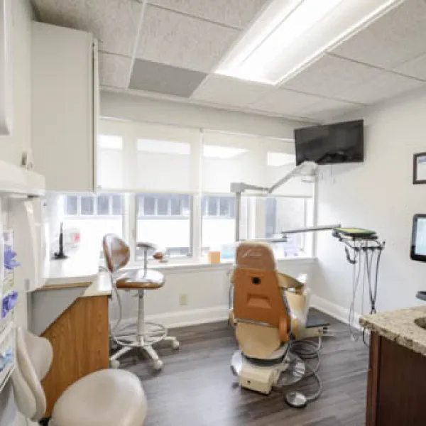 Bright dental office with patient chair, monitor displaying dental scan, stools, and large windows providing natural light