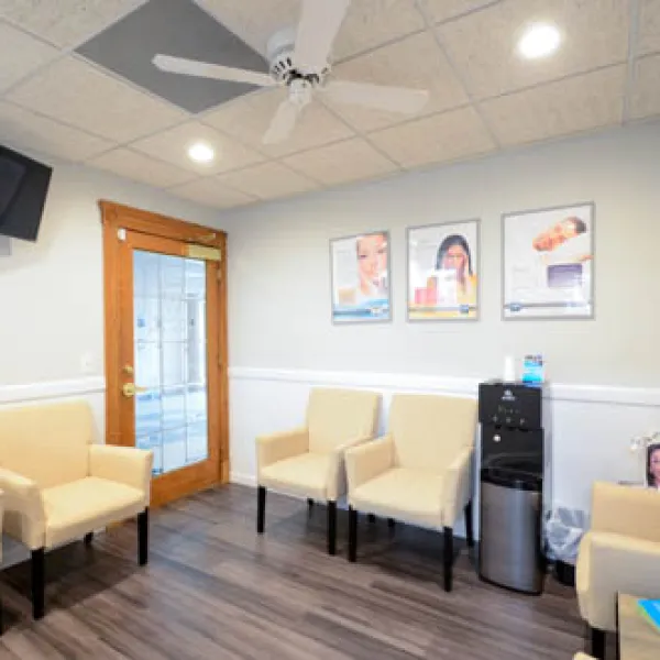 Bright waiting room with beige chairs, wooden door, water cooler, ceiling fan, and framed posters on light walls.