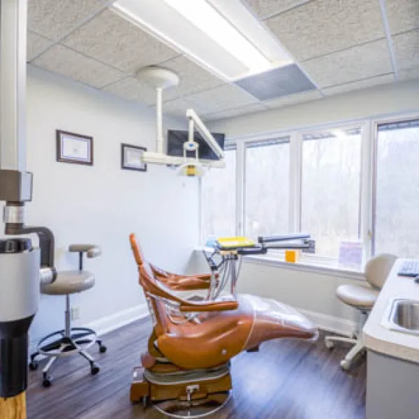 Modern dental clinic room with brown dental chair, equipment, large window, and clean white walls.