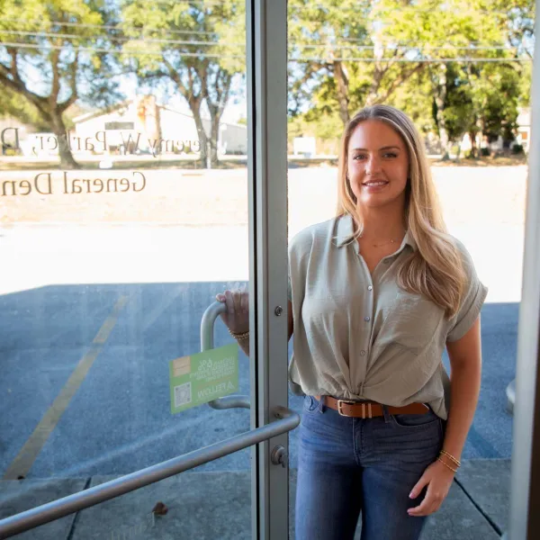 Smiling woman holding door open standing outside a dental office with trees and parking lot in background