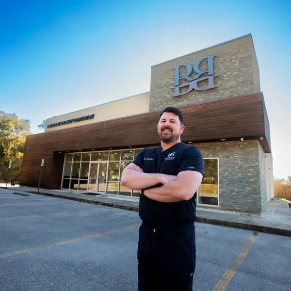 Smiling man in black scrubs stands with arms crossed outside modern dental office building under clear blue sky